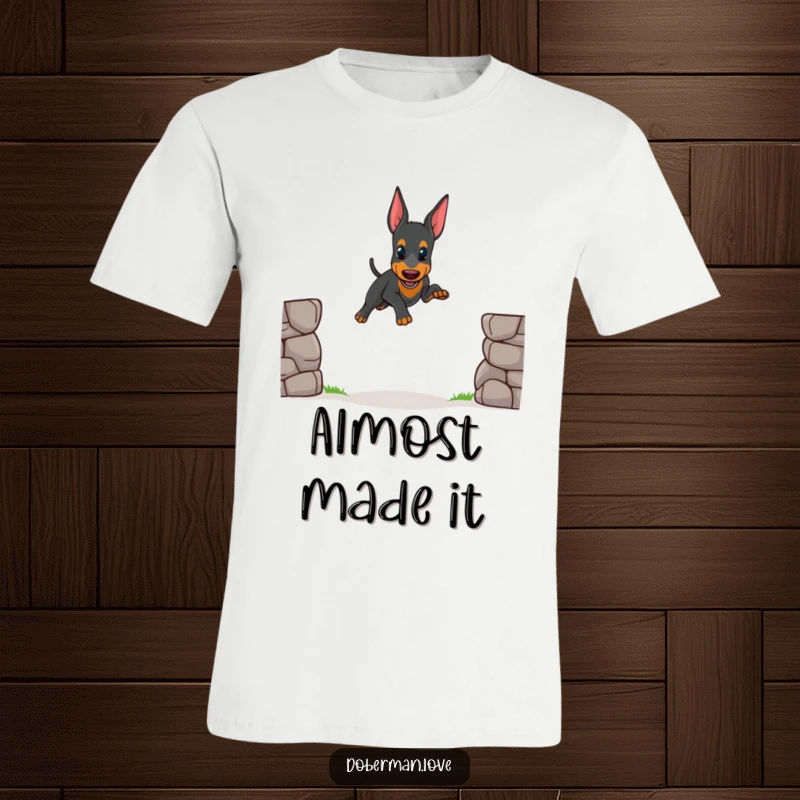 Funny t-shirt graphic of a small Doberman puppy attempting a slightly unsteady jump over a small obstacle, highlighting cute determination.